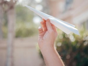 A close-up of a person's hand holding a white paper airplane outdoors, with a blurred background of greenery and buildings.